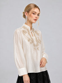 A woman wearing the Social Queen Floral Vine Embroidered Blouse in ivory, paired with a black skirt and long crystal earrings, stands against a plain gray background.