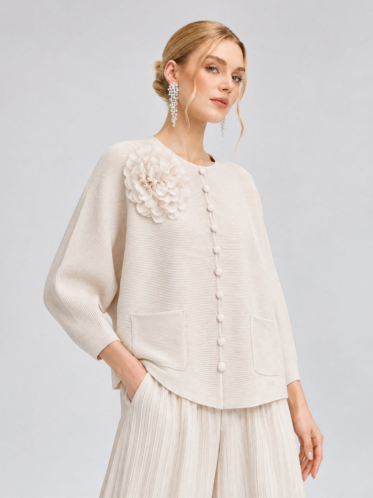 A woman models the Social Queen 3D Floral Pleated Two-Piece Set, which features cream-white pleated trousers and a textured top adorned with large buttons, two pockets, and a delicate floral appliqué on the shoulder, set against a plain light background.
