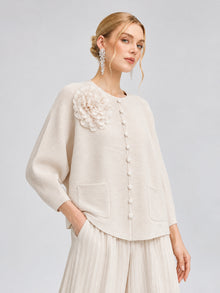 A woman models the Social Queen 3D Floral Pleated Two-Piece Set, which features cream-white pleated trousers and a textured top adorned with large buttons, two pockets, and a delicate floral appliqué on the shoulder, set against a plain light background.