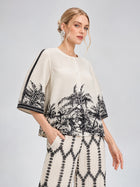 A woman models the Social Queen Palm Tree Print Pleated Blouse, a cream cropped top with a black palm tree pattern, paired with matching wide-leg pants, set against a light gray background.