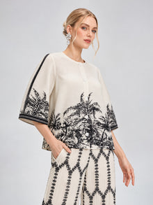 A woman models the Social Queen Palm Tree Print Pleated Blouse, a cream cropped top with a black palm tree pattern, paired with matching wide-leg pants, set against a light gray background.