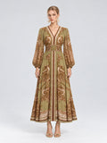 A woman wearing the Social Queen Paisley Print Mulberry Silk Dress, featuring a long, olive green and brown paisley floral pattern with long sleeves and a v-neck, stands against a plain light background. With a neutral expression and open-toed heels, she showcases this effortlessly elegant resort wear piece that combines both comfort and style.