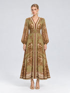 A woman wearing the Social Queen Paisley Print Mulberry Silk Dress, featuring a long, olive green and brown paisley floral pattern with long sleeves and a v-neck, stands against a plain light background. With a neutral expression and open-toed heels, she showcases this effortlessly elegant resort wear piece that combines both comfort and style.
