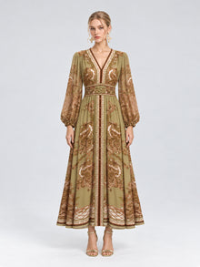 A woman wearing the Social Queen Paisley Print Mulberry Silk Dress, featuring a long, olive green and brown paisley floral pattern with long sleeves and a v-neck, stands against a plain light background. With a neutral expression and open-toed heels, she showcases this effortlessly elegant resort wear piece that combines both comfort and style.
