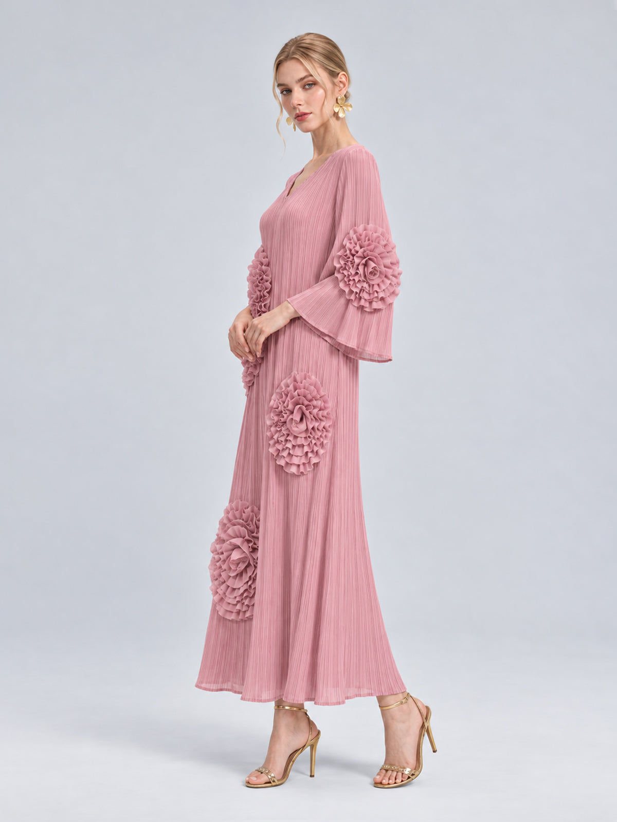 A woman stands against a plain light background wearing the Social Queen 3D Floral Pleated Dress—a pink maxi dress featuring pleats and dramatic large floral appliqué ruffle accents. She completes the look with gold heels and statement earrings.