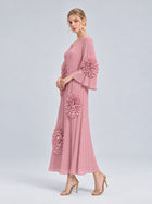 A woman stands against a plain light background wearing the Social Queen 3D Floral Pleated Dress—a pink maxi dress featuring pleats and dramatic large floral appliqué ruffle accents. She completes the look with gold heels and statement earrings.