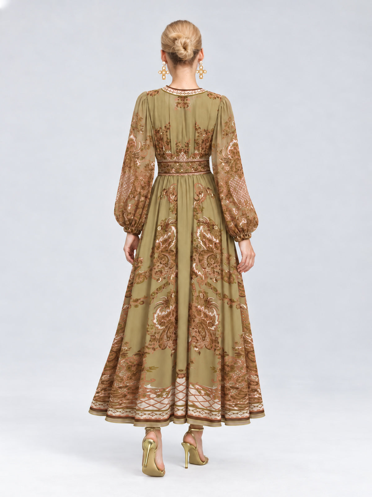 A woman stands with her back to the camera against a plain light gray background, wearing the Social Queen Paisley Print Mulberry Silk Dress—an olive green, long-sleeve, ankle-length maxi dress featuring a brown paisley floral pattern—paired with gold high-heeled sandals.