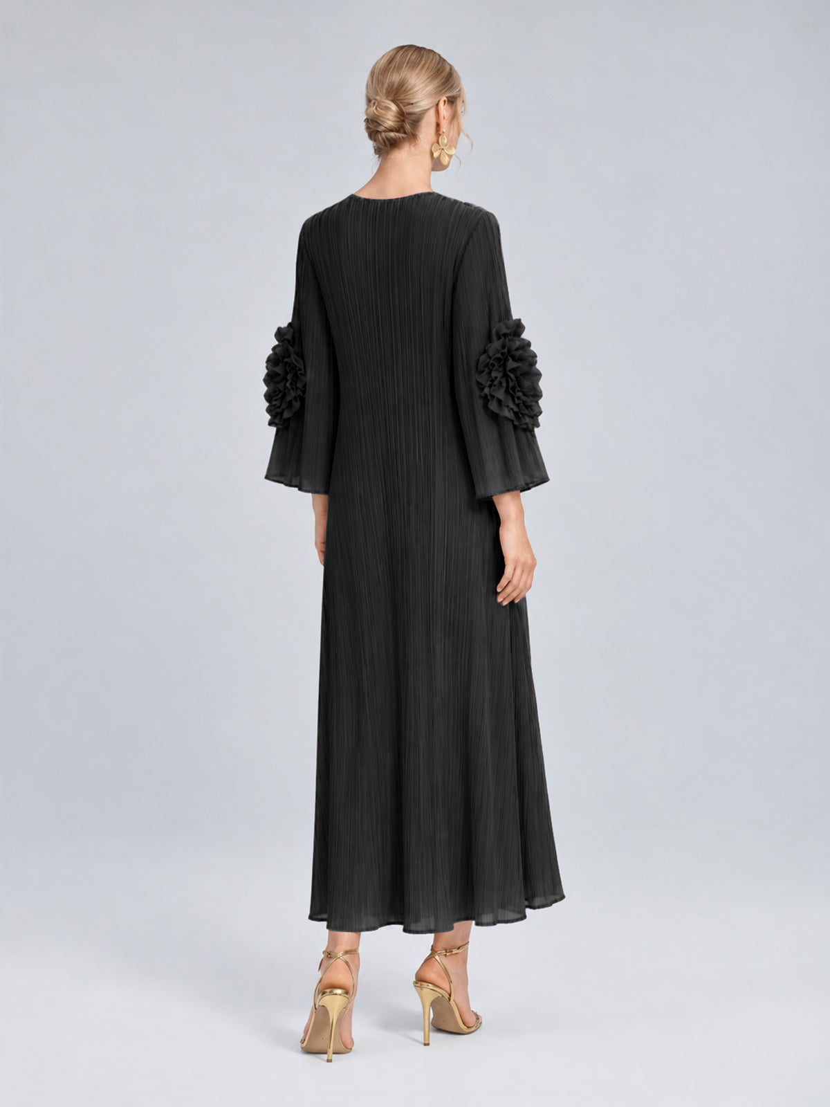 A woman stands with her back to the camera wearing the Social Queen 3D Floral Pleated Dress, a long black maxi design featuring ruffled sleeves, paired with gold earrings and gold heels.