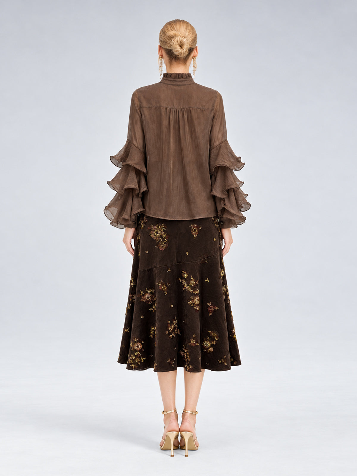 A woman stands with her back to the camera, dressed in the Social Queen Pleated Layered Ruffle Blouse in brown, crafted from soft polyester chiffon, paired with a dark floral midi skirt and beige heels against a plain light background.