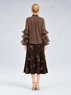 A woman stands with her back to the camera, dressed in the Social Queen Pleated Layered Ruffle Blouse in brown, crafted from soft polyester chiffon, paired with a dark floral midi skirt and beige heels against a plain light background.