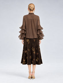 A woman stands with her back to the camera, dressed in the Social Queen Pleated Layered Ruffle Blouse in brown, crafted from soft polyester chiffon, paired with a dark floral midi skirt and beige heels against a plain light background.