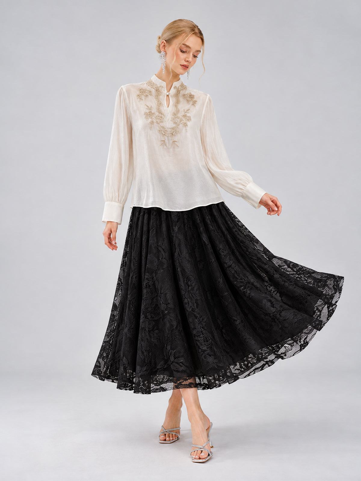A woman wears the Floral Vine Embroidered Blouse by Social Queen—an ivory shirt featuring delicate floral vine embroidery—paired with a long black lace skirt. She stands against a plain background, looking downward while holding her skirt.