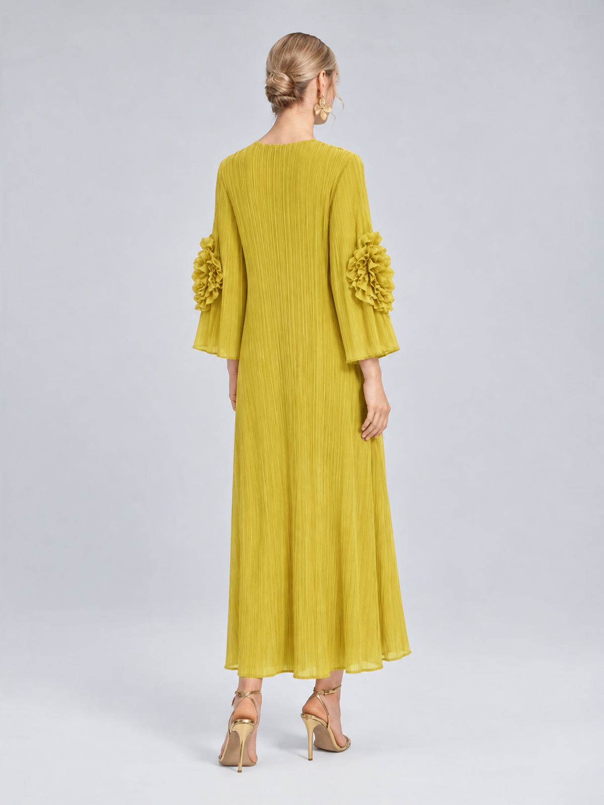 A woman stands facing away, wearing the Social Queen 3D Floral Pleated Dress, featuring pleated fabric and floral appliqué accents on the sleeves, paired with gold high heels.