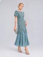 A woman poses against a plain gray background wearing the Social Queen Beaded Floral Silk Velvet Dress in light blue, featuring puff sleeves and beaded floral embroidery, styled with silver heels and a small silver clutch.