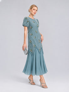 A woman poses against a plain gray background wearing the Social Queen Beaded Floral Silk Velvet Dress in light blue, featuring puff sleeves and beaded floral embroidery, styled with silver heels and a small silver clutch.