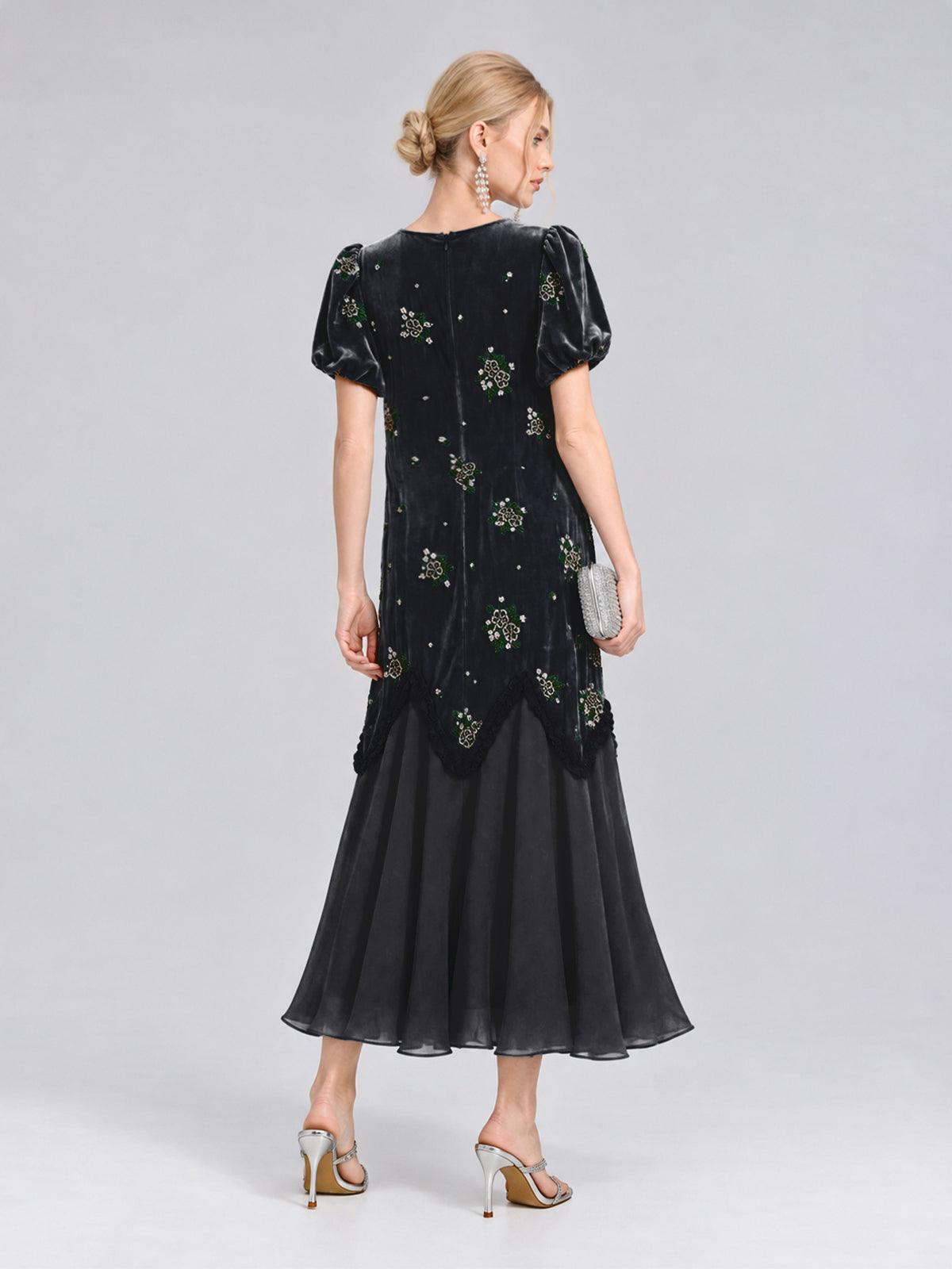 A woman stands in profile against a plain light grey background, wearing the Social Queen Beaded Floral Silk Velvet Dress—an ankle-length black dress featuring silk-blend velvet and exquisite beaded floral embroidery. She pairs the look with metallic high heels and a silver clutch.