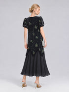 A woman stands in profile against a plain light grey background, wearing the Social Queen Beaded Floral Silk Velvet Dress—an ankle-length black dress featuring silk-blend velvet and exquisite beaded floral embroidery. She pairs the look with metallic high heels and a silver clutch.