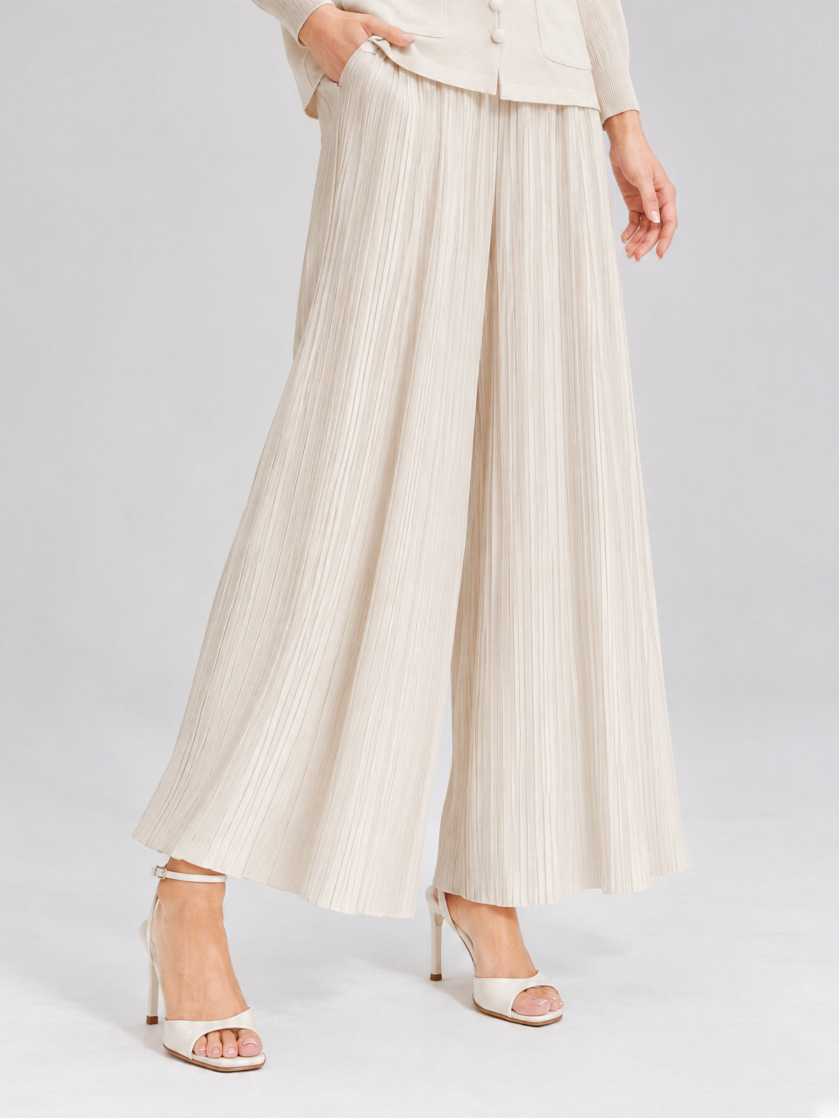 A model showcases the 3D Floral Pleated Two-Piece Set by Social Queen, featuring pleated wide-leg beige trousers and white high-heeled sandals, standing against a plain background. The ensemble captures effortless elegance, with the pleated design adding soft movement to the look.