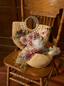 Two Social Queen 3D Dahlia Shell Woven Tote Bags sit on a carved wooden chair, with their floral appliqué and shell details standing out against a wood-paneled background.