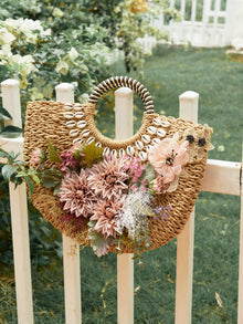 The Social Queen 3D Dahlia Shell Woven Tote Bag, featuring shell accents, pink flowers, and green leaves, hangs on a white wooden garden fence.