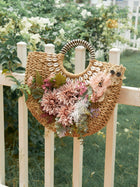 The Social Queen 3D Dahlia Shell Woven Tote Bag, featuring shell accents and artificial flowers, hangs on a white wooden fence in a garden.