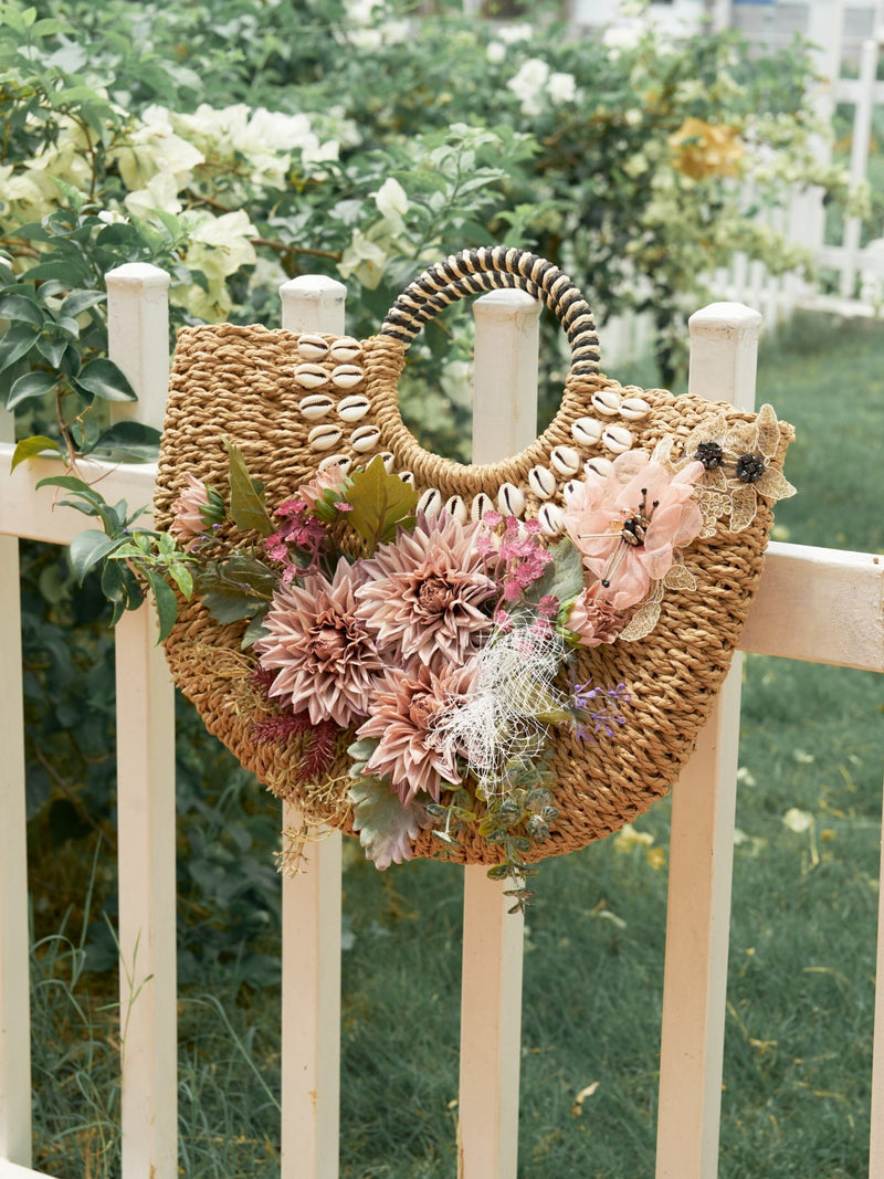 The Social Queen 3D Dahlia Shell Woven Tote Bag, featuring shell accents and artificial flowers, hangs on a white wooden fence in a garden.