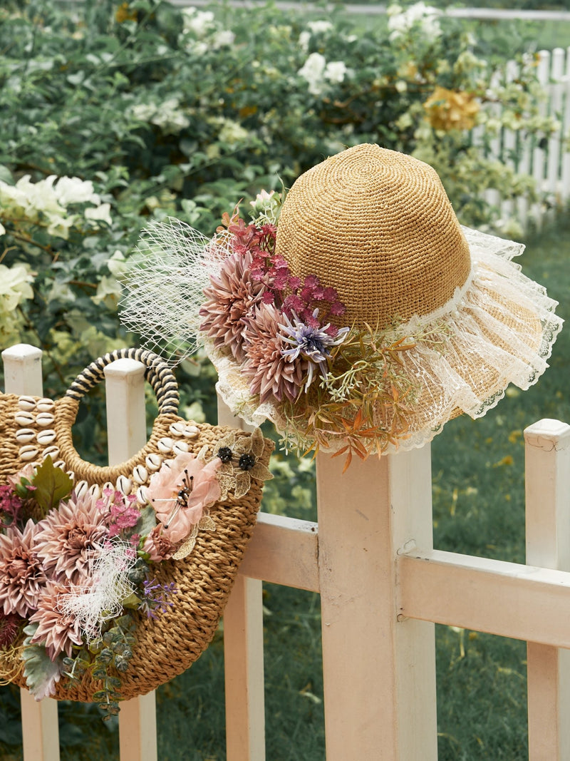 A straw hat with lace, netting, and faux flowers sits on a white fence beside the Social Queen 3D Dahlia Shell Woven Tote Bag featuring delicate floral accents.