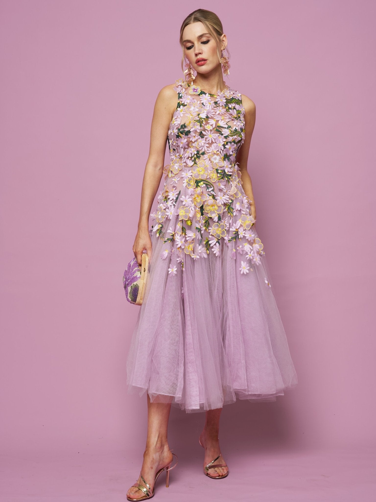 A woman stands against a pink background wearing the Social Queen 3D Daisy Embroidered Tulle Dress, styled sleeveless with floral details, paired with a small handbag and high-heeled sandals.