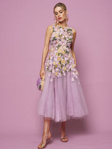 A woman stands against a pink background wearing the Social Queen 3D Daisy Embroidered Tulle Dress, styled sleeveless with floral details, paired with a small handbag and high-heeled sandals.