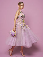 A woman wears the Social Queen 3D Daisy Embroidered Tulle Dress, a sleeveless lavender A-line with hand-sewn daisy details, holding a matching clutch and gold heels, standing against a pink background.