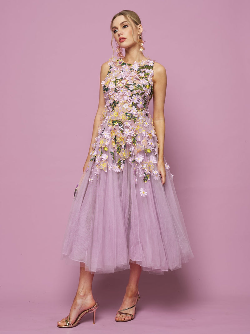 A woman stands against a lilac background in the Social Queen 3D Daisy Embroidered Tulle Dress, an A-line silhouette with sleeveless design and daisy details, paired with heeled sandals.