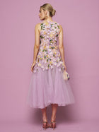 A woman faces away from the camera against a pink background, wearing Social Queen’s 3D Daisy Embroidered Tulle Dress with pink heels and a small clutch. The sleeveless, lilac mid-length dress features hand-embroidered 3D daisies.
