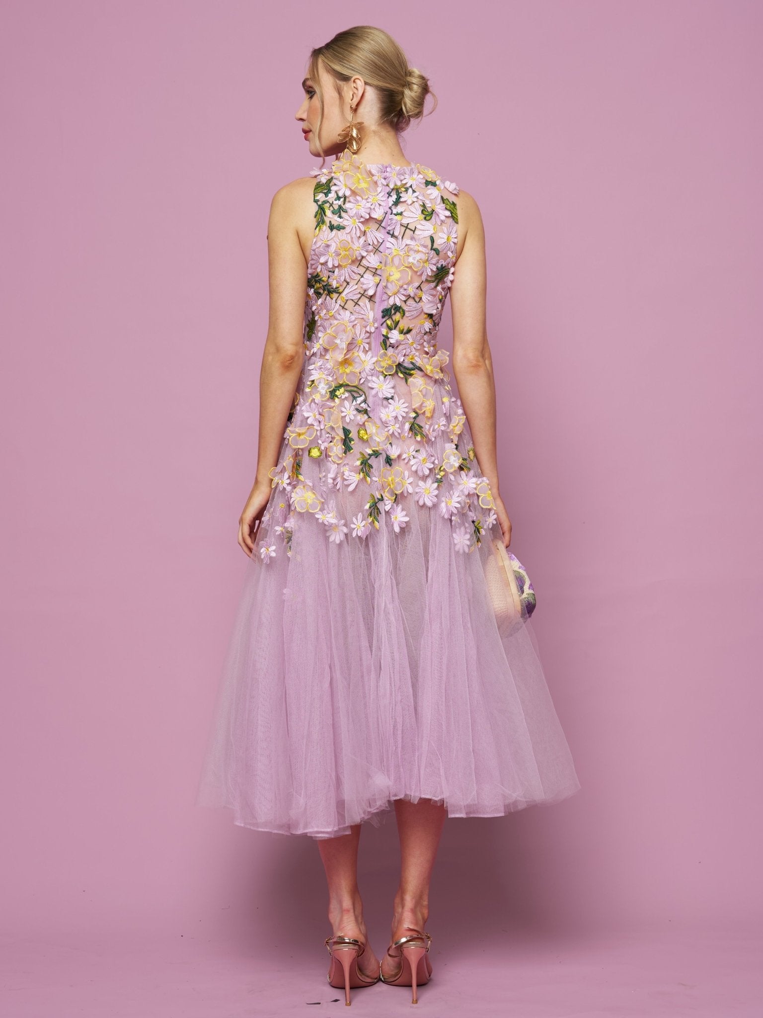 A woman faces away from the camera against a pink background, wearing Social Queen’s 3D Daisy Embroidered Tulle Dress with pink heels and a small clutch. The sleeveless, lilac mid-length dress features hand-embroidered 3D daisies.