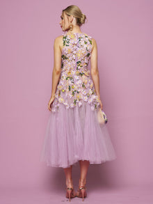 A woman faces away from the camera against a pink background, wearing Social Queen’s 3D Daisy Embroidered Tulle Dress with pink heels and a small clutch. The sleeveless, lilac mid-length dress features hand-embroidered 3D daisies.