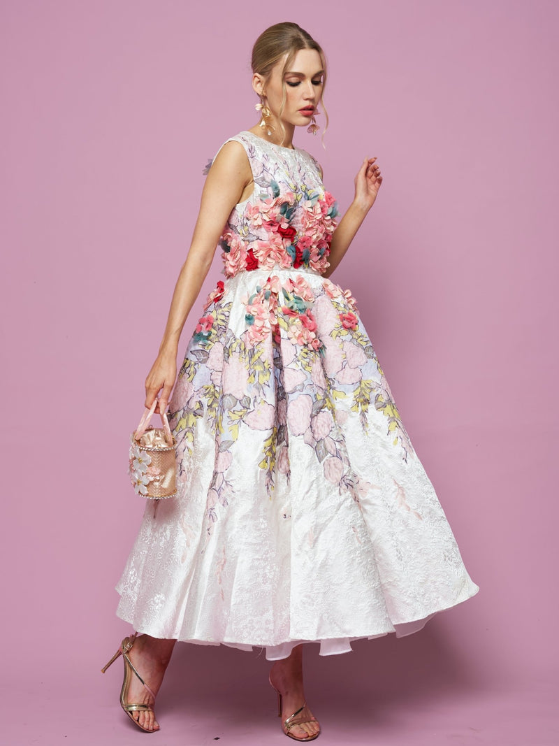 A woman poses against a pink background in the Social Queen 3D Floral Embellished Jacquard Gown, paired with a small purse and gold heels.