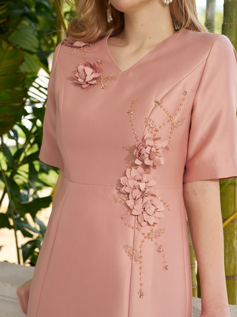 A woman wears the Social Queen 3D Floral Embellished Satin Dress, featuring a blush pink mermaid silhouette, V-neck, and 3D floral appliqués on the chest and side, standing before lush green plants.