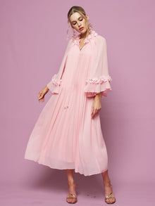 A woman models the Social Queen 3D Floral Embellished Silk Dress, featuring light pink mulberry silk, pleats, long sleeves, and floral neckline accents, set against a solid pink background.
