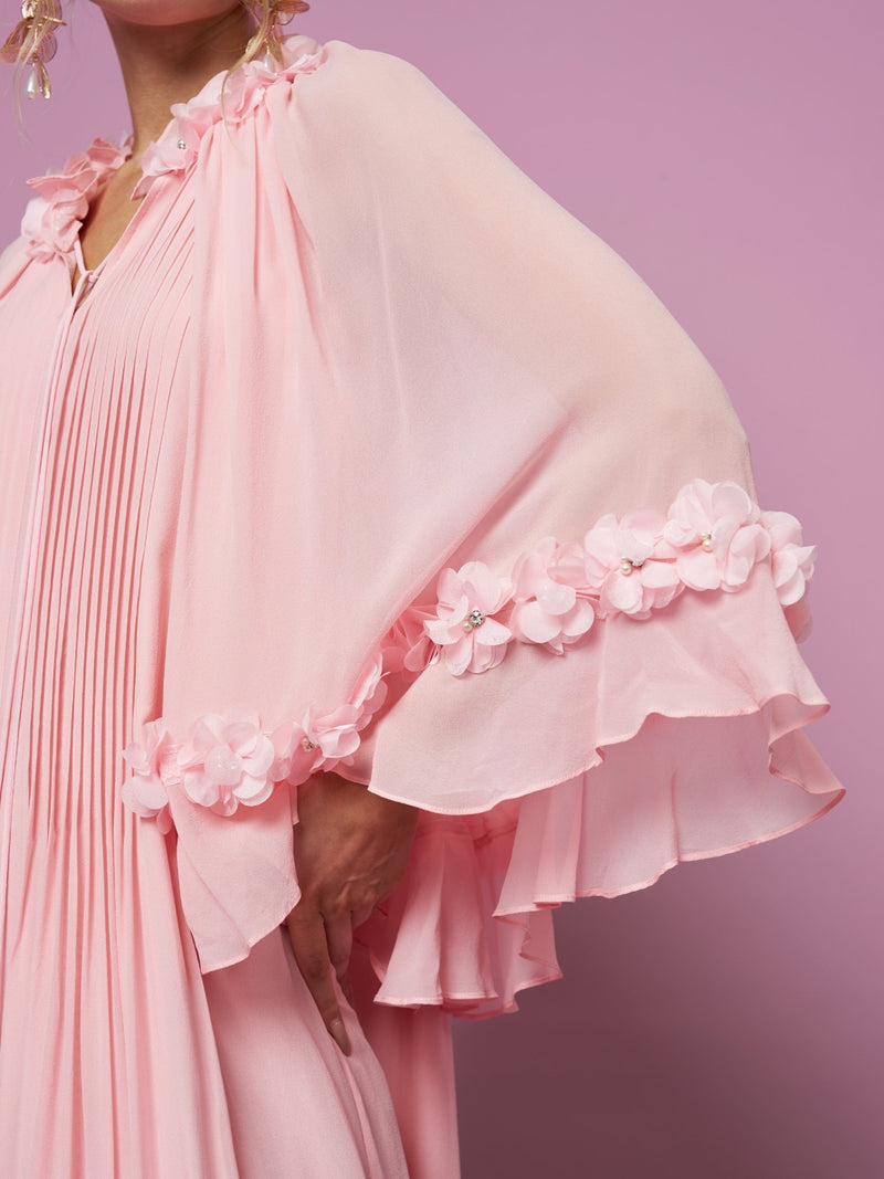 A person stands against a solid pink background wearing Social Queen's 3D Floral Embellished Silk Dress, featuring flowing light pink Mulberry silk, pleated fabric, and ruffled sleeves trimmed with 3D floral appliqués.