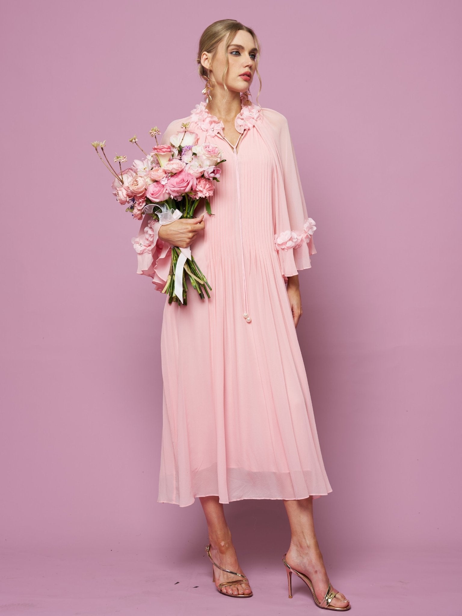 Woman in a light pink Social Queen 3D Floral Embellished Silk Dress with long sleeves and 3D floral appliqué, holding a bouquet of pink flowers against a pink background, styled with heeled sandals.