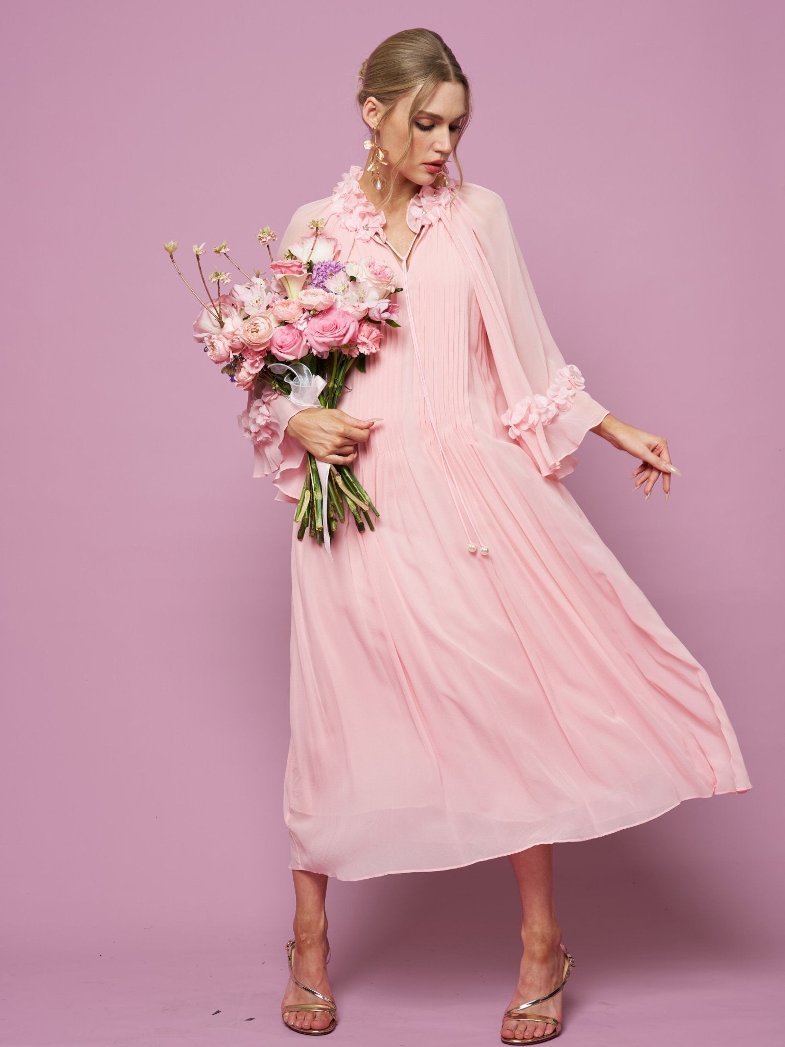 A woman wears Social Queen’s 3D Floral Embellished Silk Dress, light pink and adorned with floral appliqué, as she holds a bouquet of pink and white flowers against a pink background.