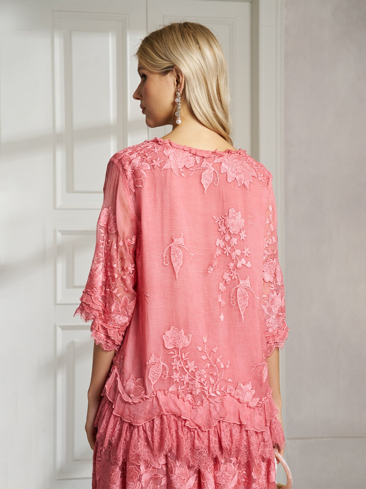 A woman with long blonde hair, seen from behind indoors, wears the Social Queen 3D Floral Embroidered 100% Silk Ruffle Blouse in pink.