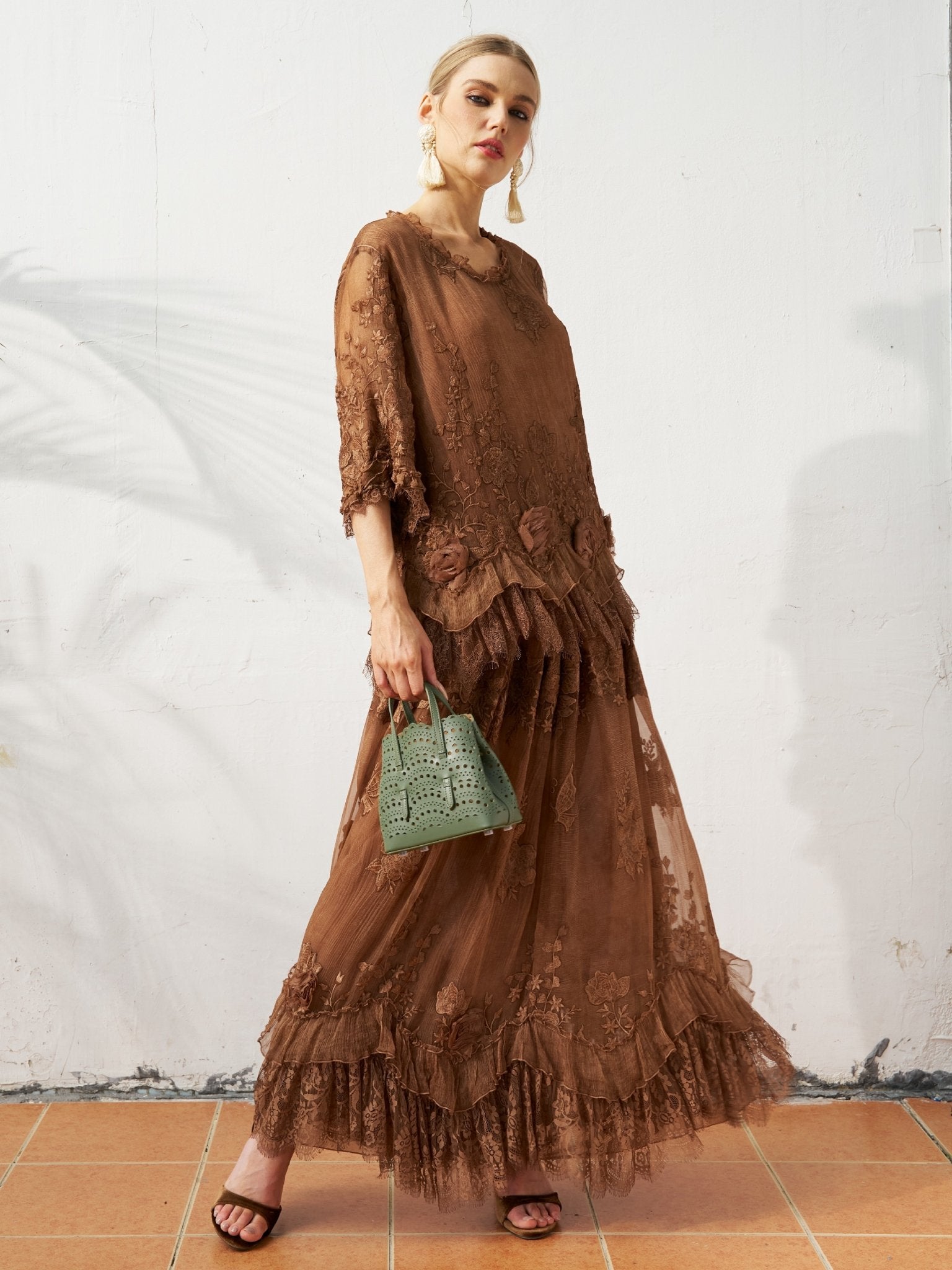A woman stands against a white wall, wearing the Social Queen 3D Floral Embroidered 100% Silk Ruffle Blouse with black sandals and holding a small green handbag.