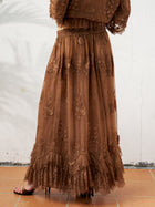 A person wears the Social Queen 3D Floral Embroidered 100% Silk Ruffle Skirt, featuring brown silk chiffon, floral embroidery, and a ruffled hem, while standing on a tiled floor against a white wall.