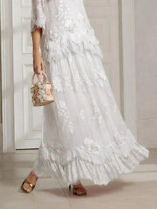 A woman stands indoors near a white door wearing the Social Queen 3D Floral Embroidered 100% Silk Ruffle Skirt, paired with a small pink floral handbag and gold open-toe heels.