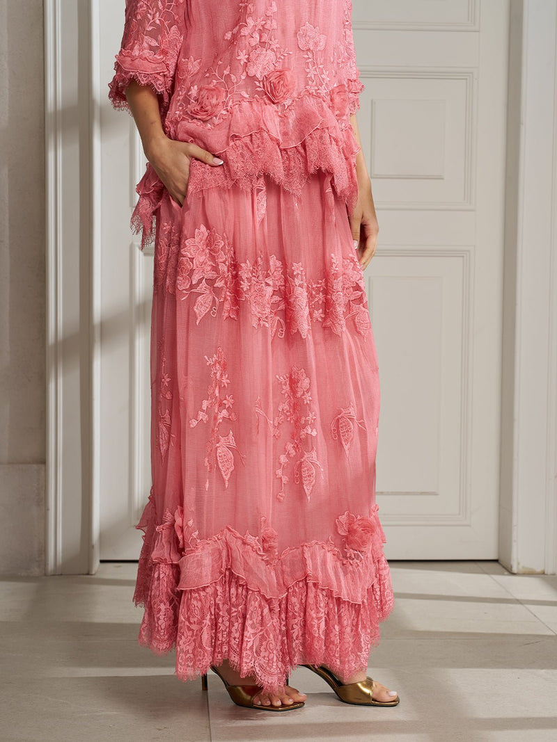 Indoors, a person models the Social Queen 3D Floral Embroidered 100% Silk Ruffle Skirt in pink, featuring ruffled details and 3D floral accents, styled with gold open-toe heels.