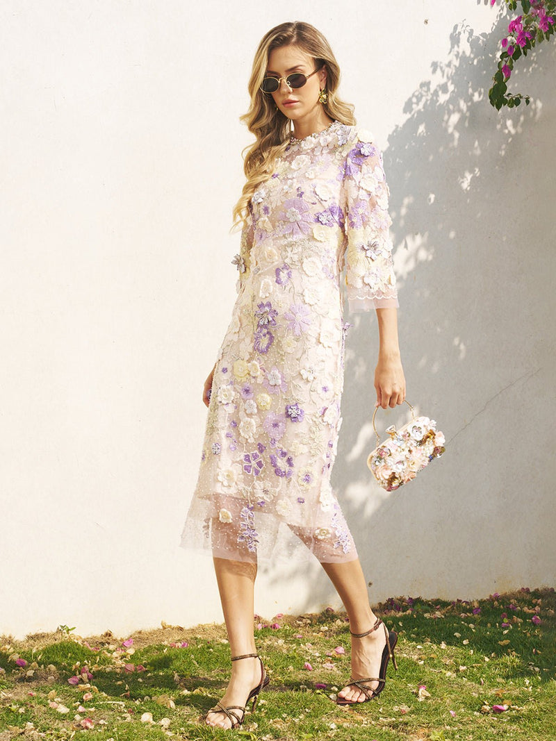 A woman stands outdoors on grass near a white wall, wearing the Social Queen 3D Floral Embroidered Beaded Dress and holding a matching handbag, with small flowers scattered on the ground nearby.
