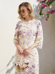 A woman in the Social Queen 3D Floral Embroidered Beaded Dress and sunglasses stands against a light wall, with purple flowers and greenery in the corner.