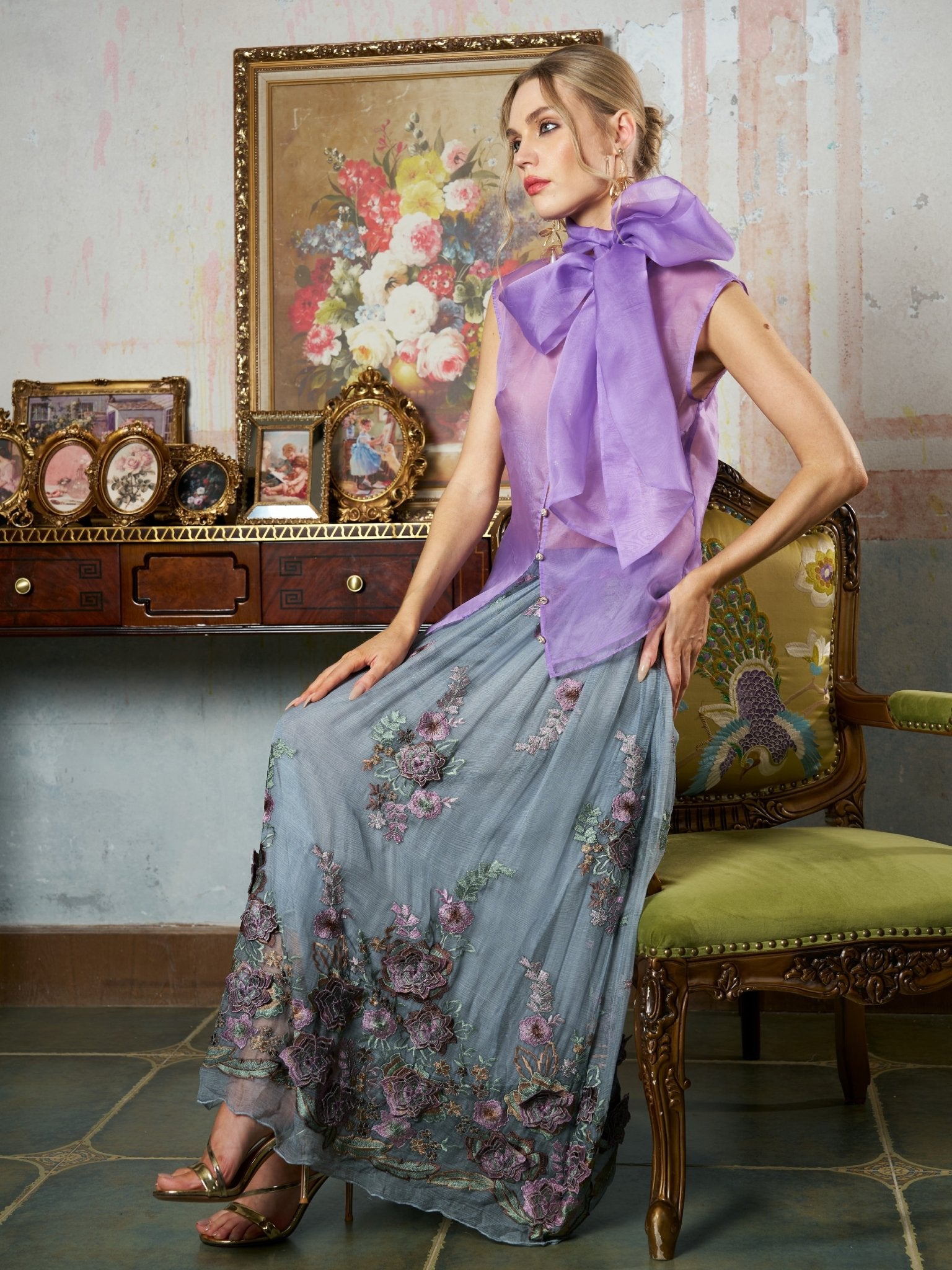 A woman in a lavender blouse and the Social Queen 3D Floral Embroidered Silk Skirt sits on a green chair by a wooden desk with framed photos, set against a backdrop with a floral painting.