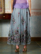A person stands indoors wearing the Social Queen 3D Floral Embroidered Silk Skirt with sheer lavender and gray florals, paired with a purple top and gold high-heeled sandals.