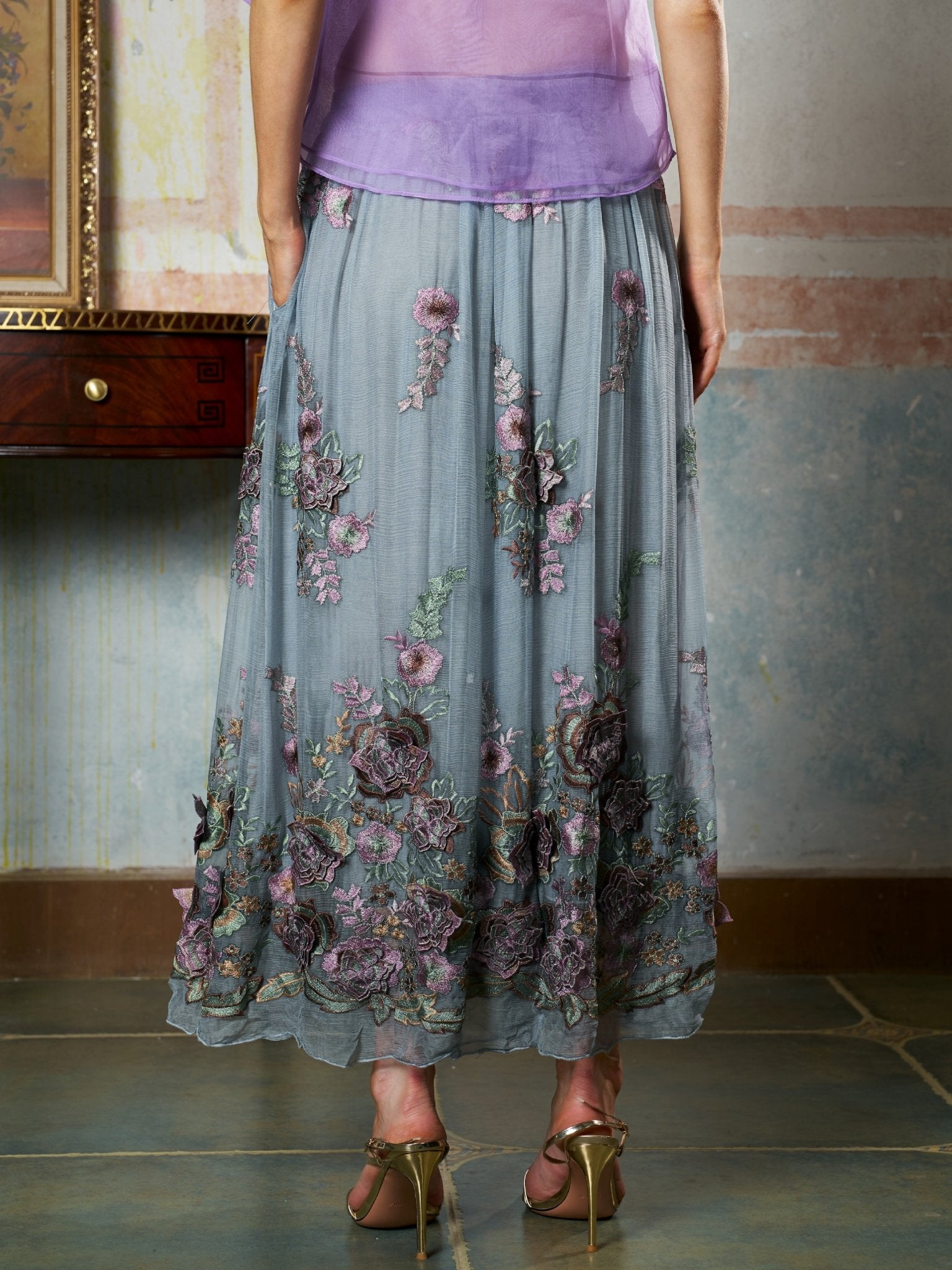 A person stands indoors wearing the Social Queen 3D Floral Embroidered Silk Skirt with sheer lavender and gray florals, paired with a purple top and gold high-heeled sandals.
